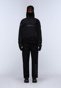 Persona vestita con abbigliamento invernale nero, inclusi giacca con cappuccio, guanti, pantaloni, stivali e occhiali con lenti arancioni, su uno sfondo chiaro e neutro.
