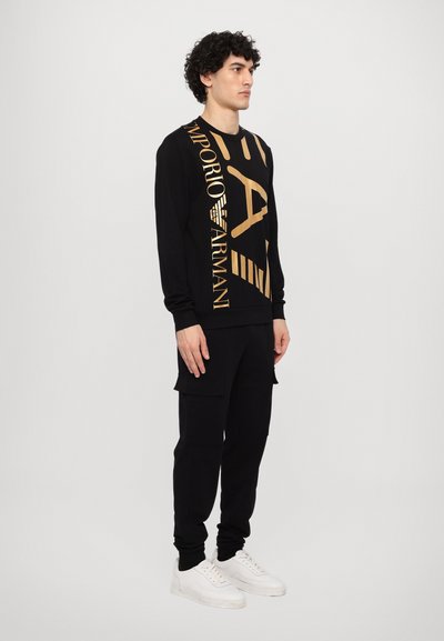 Sweatshirt noir avec des détails de logo doré, associé à un pantalon cargo noir. Le modèle porte des baskets blanches, présentant une tenue décontractée.