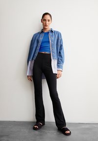 Een spijkerblouse over een blauwe crop top, gecombineerd met zwarte flared broeken en donkere slidesandalen. De blouse heeft knopen en strepen.