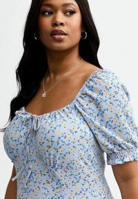 Robe bleu clair à motifs floraux avec des manches bouffantes et un décolleté carré. Tissu doux et texturé avec un cordon de serrage à l'avant.