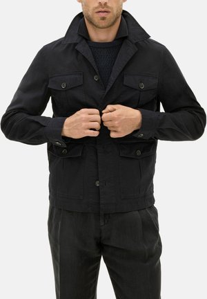 Homme boutonnant une veste noire à quatre poches à rabat, portant un pull texturé noir et un pantalon noir, debout devant un fond blanc.