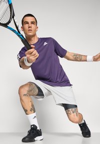 Camiseta deportiva morada, pantalones cortos grises con detalles en negro, zapatillas de tenis negras, muñequeras, sosteniendo una raqueta de tenis, mostrando tatuajes en la pierna.