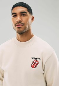 Lyse beige sweatshirt med rund halsudskæring, prydet med et rødt og sort broderet Rolling Stones-logo på venstre brystområde.