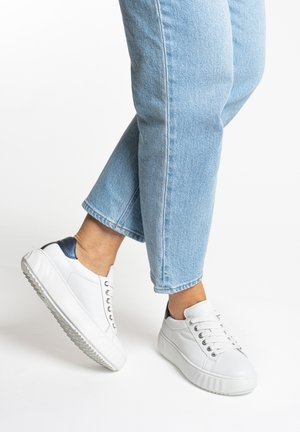 Person wearing light blue jeans and white sneakers with dark blue heel accents, posing with one foot slightly lifted.