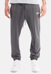 Graue Jogginghose mit elastischen Bündchen, Kordelzugbund, vorderen Taschen und einem kleinen Logopatch auf der linken Seite. Kombiniert mit weißen Sneakers.