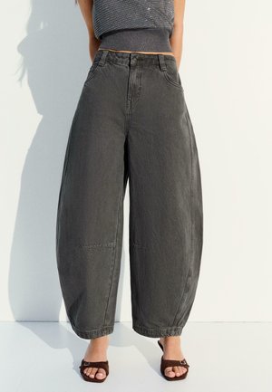 Kobieta w szerokich spodniach o wysokim stanie w kolorze węgla, szarym dzianinowym topie bez rękawów i brązowych sandałach na otwarte palce, stojąca przy białej ścianie.