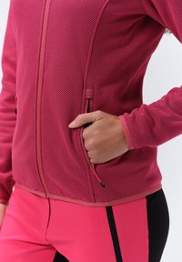 Rosa strukturierte Jacke mit Reißverschluss-Taschen und kontrastierenden Details, kombiniert mit rosa und schwarzer, figurschmeichelnder Hose. Sichtbares Wabenmuster.