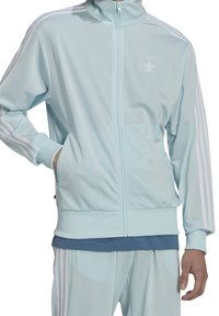 Lichtblauw jack met ritssluiting van gladde stof. Voorzien van witte gestreepte accenten op de mouwen en een logo op de borst. Zakjes aan de zijkanten.