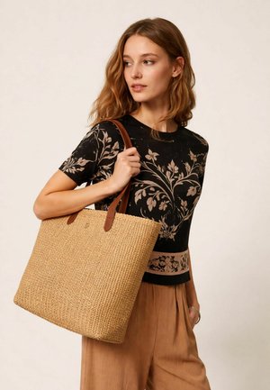 Donna che tiene una grande borsa a tracolla intrecciata con manici in pelle marrone, indossa una maglietta nera a fiori e pantaloni plissettati color beige, guarda di lato.