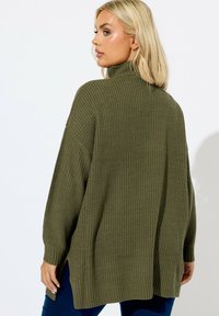Olivgrüner Strickpullover mit hohem Kragen, überschnittenen Schultern und seitlichen Schlitzen. Texturierter Stoff mit gerippten Bündchen und lockerer Passform.