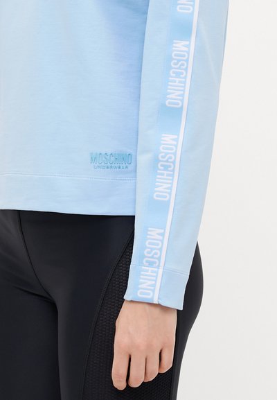 Light blue long-sleeve top with "Moschino" logo printed along the sleeve. Fabric appears smooth; features ribbed cuff and hem.