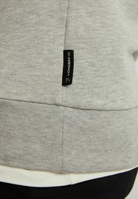 Felpa grigia con trama a coste, caratterizzata da un'etichetta nera sul lato. Indossata sopra una camicia bianca, con tessuto nero sul fondo.