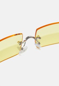 Ochelari de soare cu tentă galbenă, cadre portocalii unghiulare, accente metalice și suporturi pentru nas transparente, având un design minimalist și geometric.