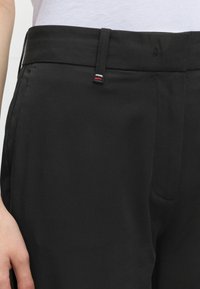 Gros plan d'une personne portant un pantalon noir avec des passants de ceinture, l'un des passants présente de petites rayures blanches, rouges et noires, avec une chemise blanche rentrée.