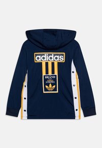 Sudadera azul marino con un gran logotipo amarillo de Adidas y tres franjas verticales. Presenta paneles laterales blancos con detalle de botones. Tejido suave.