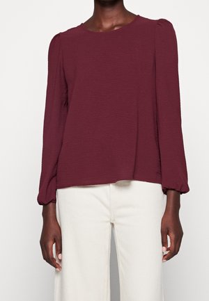 Vrouw draagt een lange mouwen gestructureerde bordeauxblouse met opgeblazen schouders en een witte broek tegen een eenvoudige lichte achtergrond.