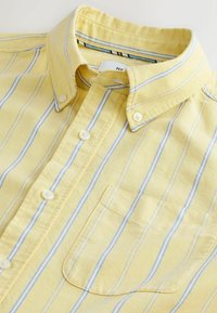 Žltá košeľa na zapínanie s modrými a bielymi vertikálnymi pruhmi, predným vreckom a golierom na gombíky, rozložená na rovnej ploche.