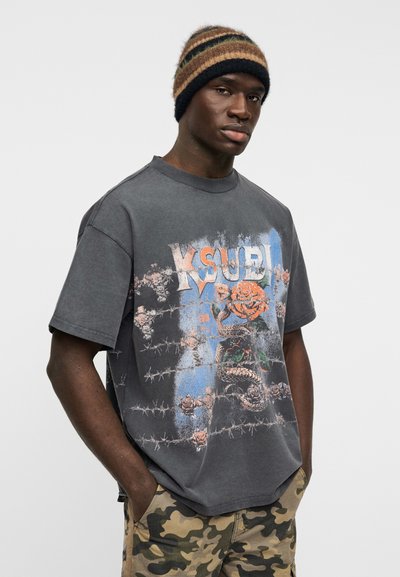 Γκρι φαρδιά μπλούζα με εμπρός σχέδιο που απεικονίζει τριαντάφυλλα, συρματόπλεγμα και φίδι, συνδυασμένη με ριγέ πλεκτό σκουφάκι και κοντά παντελόνια καμουφλάζ.