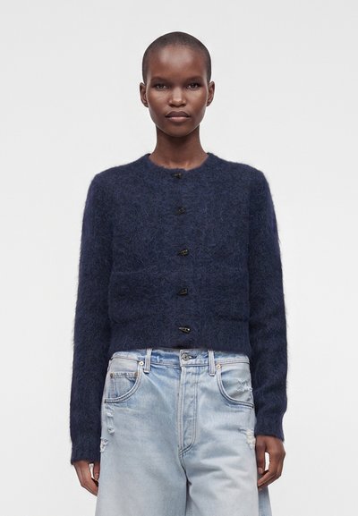 Personne portant un cardigan bleu marine duveteux boutonné et un jean taille haute bleu clair avec des détails usés, debout devant un fond uni.