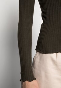 Haut à manches longues côtelé vert olive avec un ourlet festonné, associé à un tissu de couleur claire. Texture douce, coupe ajustée.