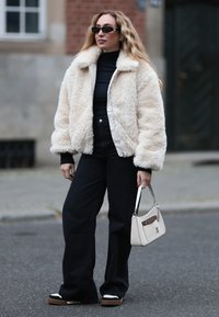 Jakna bomber iz kremaste umetne dlake, črn pulover z visokim ovratnikom, črne jeans hlače z visokim pasom in črni športni copati z belimi poudarki. V roki drži belo torbico.