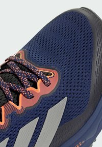 Sininen urheilujalkine, jossa on hengittävä verkkomateriaali, teksturoidut mustat yksityiskohdat, oranssit yksityiskohdat ja näyttävät valkoiset raidat sivuilla.