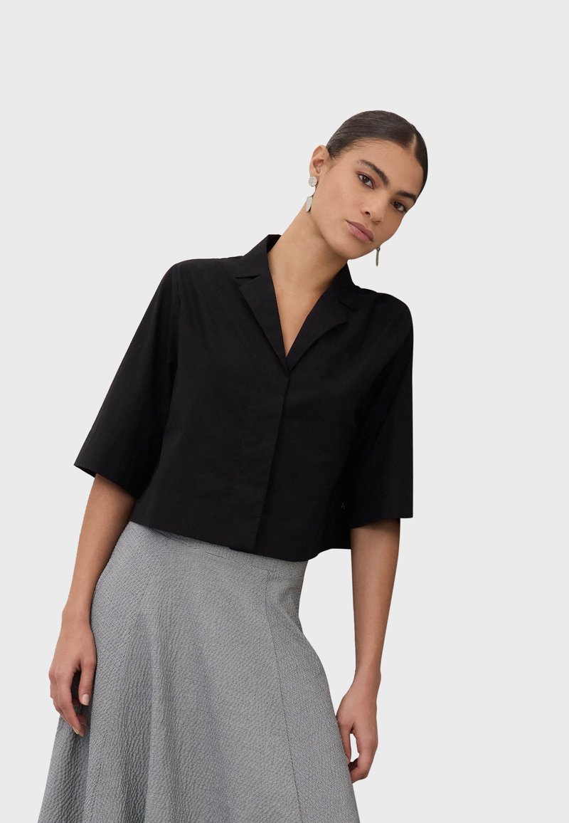 Mujer con una camisa negra de manga corta con cuello y una falda gris texturizada de cintura alta, posando contra un fondo claro y liso.