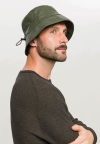 Cappello a secchiello impermeabile verde con una larga visiera, superficie texturizzata e cordino regolabile sul lato. Indossato da un uomo con un maglione scuro.