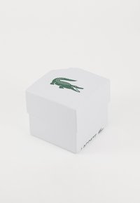 Caixa de cartão branca com uma superfície texturizada, apresentando um logotipo de crocodilo verde na tampa e o nome da marca do lado.