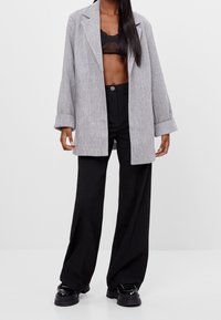 Grauer Fischgrätenmuster-Übergröße-Blazer, schwarzes Mesh-Bralette darunter, kombiniert mit schwarzen, weit geschnittenen Hosen und Plateauschuhen. Glatter Stoff, lockere Passform.