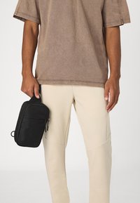 Schwarze Stoff-Crossbody-Tasche mit Reißverschlusstaschen, wird in einer Hand gehalten. Beige Hose mit gestickten Details, kombiniert mit einem muted braunen übergroßen T-Shirt.
