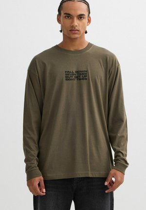 Camisa de manga larga verde oliva hecha de una tela suave que presenta un texto motivacional impreso en negro en el centro. Ajuste simple y relajado.