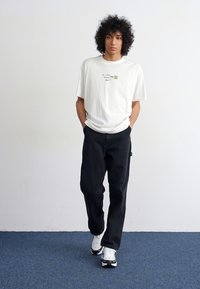 Hvid t-shirt med korte ærmer og grøn grafisk design, kombineret med sorte løse bukser og hvide sneakers, stående på blå tæppe.