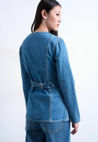 Femme portant une veste en denim bleu ajustée avec un détail ceinturé dans le dos, assortie à un jean en denim bleu, regardant sur le côté.