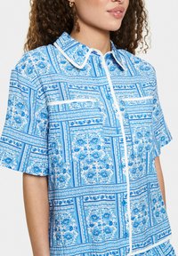 Košeľa s krátkym rukávom z ľahkého materiálu, s modro-bielym kvetinovým vzorom, s gombíkmi na prednej strane a kontrastným golierom a lemovaním vrecka.