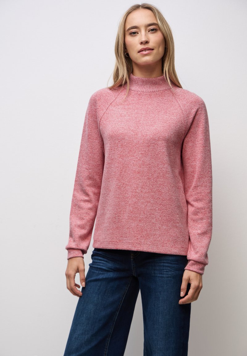 Rosa gestrickter Pullover mit hohem Kragen, langen Ärmeln und lockerer Passform, kombiniert mit dunkelblauen Weitbein-Jeans. Weiche Textur, minimalistisches Muster.