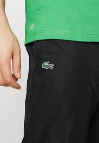 Grön kortärmad skjorta med "ULTRA DRY"-etikett, parat med svarta shorts som har en liten grön och röd krokodil-logotyp på sidan.