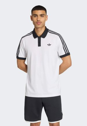 Mann trägt weißes Adidas-Poloshirt mit schwarzem Kragen und Streifen, kombiniert mit schwarzen Shorts, steht mit den Händen hinter dem Rücken.