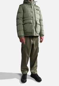 Olivgrüne Steppjacke mit Kapuze, ausgestattet mit einem Logo-Patch und Druckknöpfen, kombiniert mit olivfarbenen Cargo-Hosen und schwarzen Sneakers.