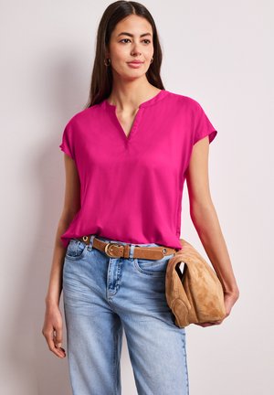 Femme portant un chemisier rose vif à col en V, jean bleu clair avec ceinture marron, tenant une veste en daim beige, debout contre un mur clair uni.