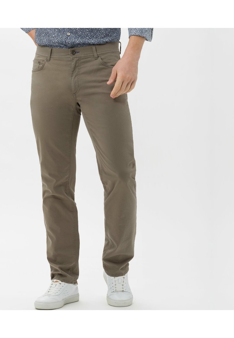Homme portant un pantalon chino marron clair, des baskets blanches et une chemise bleue à motifs, debout devant un fond blanc uni.