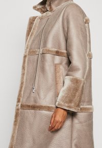 Beige faux fur coat with a textured finish, featuring a zip closure, oversized collar, and contrasting smooth panels and sleeves.