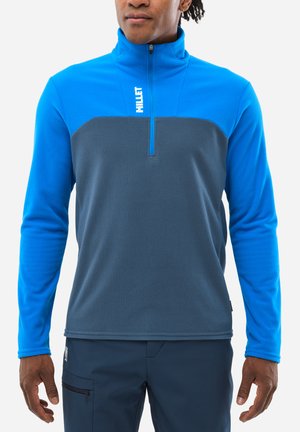 Pull en polaire bleu et gris avec demi-zip, présentant un corps texturé et des manches lisses, ainsi qu'un logo sur le haut de la poitrine. Le porteur est posé de manière décontractée.