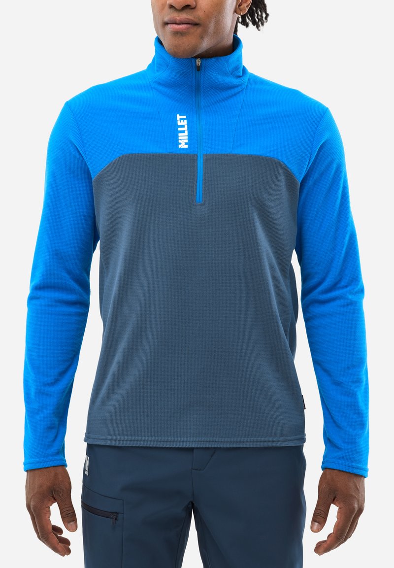 Blauer und grauer Fleece-Pullover mit halbem Reißverschluss, der einen strukturierten Körper und glatte Ärmel aufweist, sowie ein Logo auf der oberen Brust. Der Träger ist lässig posiert.