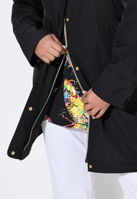 Svart zip-up jacka med guldfärgad detaljer som avslöjar en färgglad mönstrad topp under. Modellen bär vita byxor. Slät textur, avslappnad design.