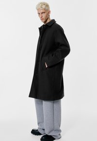 Zwarte wollen jas met een klassieke kraag, zijzakken en een rechte snit, gecombineerd met grijze joggingbroek en zwarte chunky schoenen.