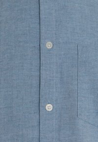 Chemise bleu clair à manches longues en tissu texturé, avec une poche plaquée, des boutons blancs et un design classique et épuré.