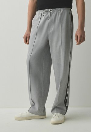 Mann trägt hellgraue weite Hose mit schwarz-weißen Seitenstreifen, kombiniert mit weißen Sneakers und einem schwarzen Oberteil.