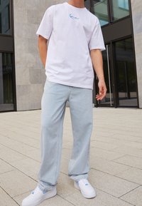 T-shirt bianca in cotone con logo ricamato blu, abbinata a pantaloni in denim oversize blu chiaro e sneakers bianche con accenti blu chiaro.