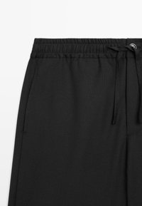 Pantalon à taille élastique noire avec cordon de serrage et poches latérales, présenté sur un fond blanc, mettant l'accent sur la partie supérieure avant.